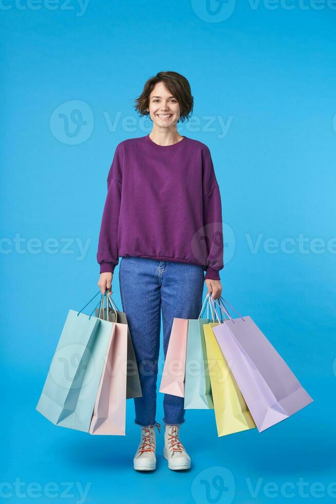 Indoor shot of young pleased dark haired curly lady being in nice mood after shopping day and holding paper bags, dressed in casual wear while posing over blue background photo