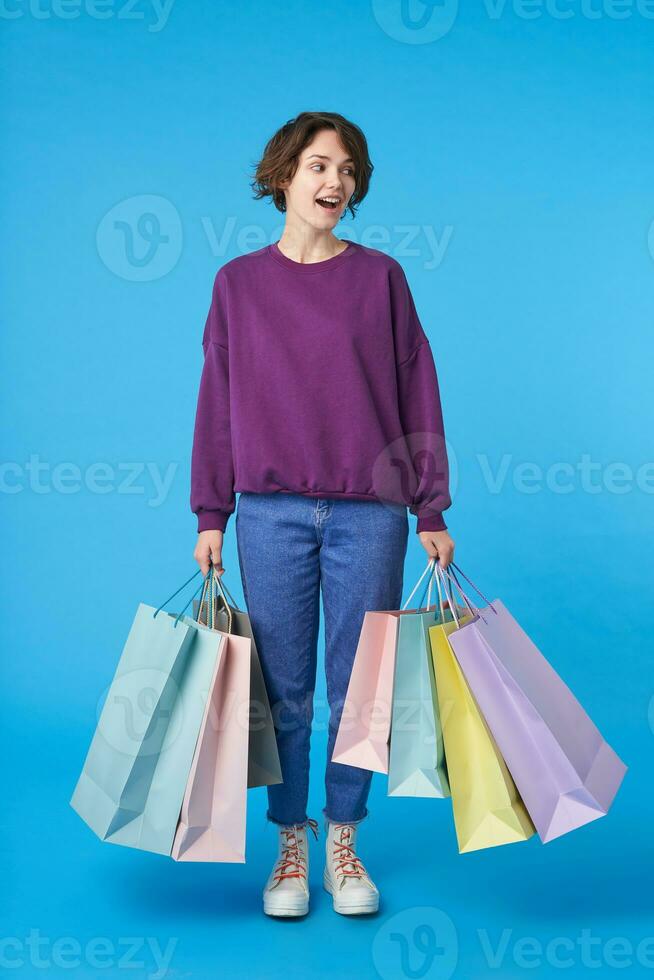 Joyful young pretty dark haired curly woman with casual hairstyle walking over blue background with colored paper bags with purchases, wearing purple sweatshirt, jeans and sneakers photo