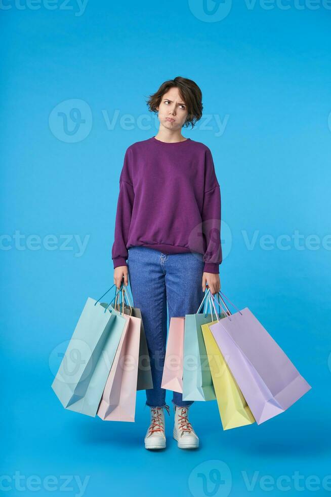 Displeased young attractive dark haired curly lady with short haircut keeping paper bags in her hands and looking tiredly upwards while standing over blue background photo