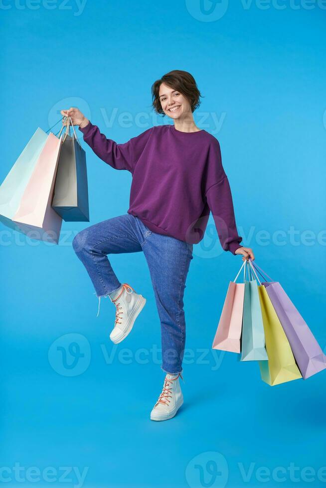 Joyful young lovely short haired curly female with casual hairstyle raising hand with purchases and looking cheerfully at camera with wide smile, isolated over blue background photo