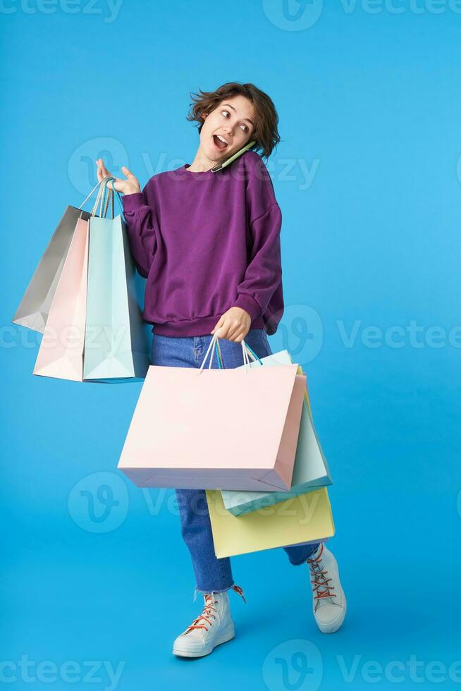 Agitated young pretty short haired curly woman dressed in casual wear answering phone call while making shopping, standing over blue background in casual clothes photo