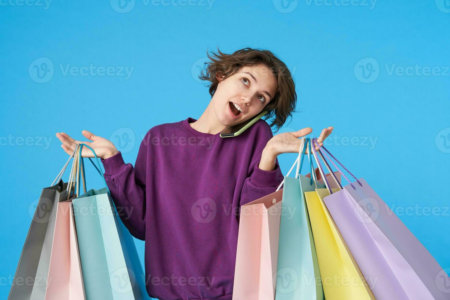 Indoor shot of young pretty curly brunette woman with short haircut making call with her smartphone while shopping, keeping paper bags in raised hands, isolated over blue background photo