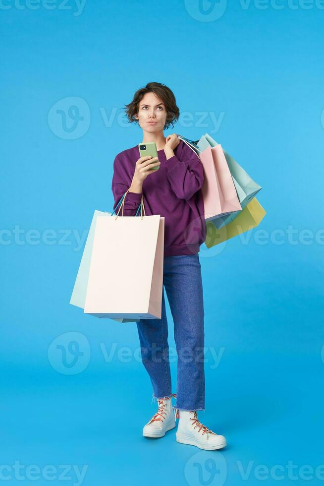 Puzzled young pretty short haired curly female keeping mobile phone in raised hand and looking pensively upwards, posing over blue background with shopping bags photo