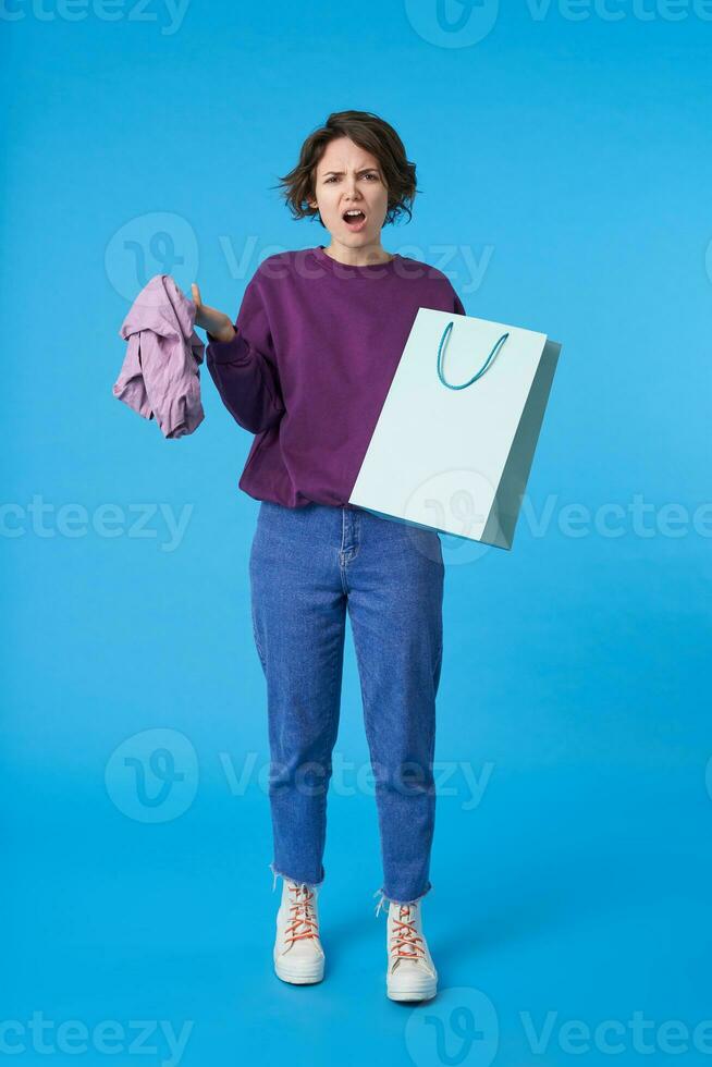 Disgruntled young lovely dark haired curly lady with natural makeup looking at camera with pout while keeping clothes and paper bag in raised hands, isolated over blue background photo