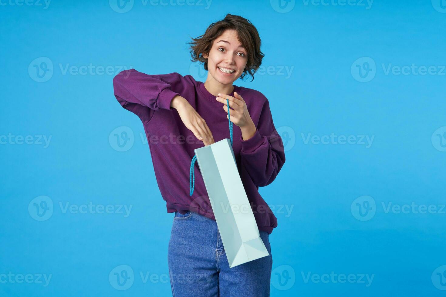Indoor photo of young cheerful curly brunette female sticking hand inside of bag and looking excitedly at camera with surprised face, standing over blue background