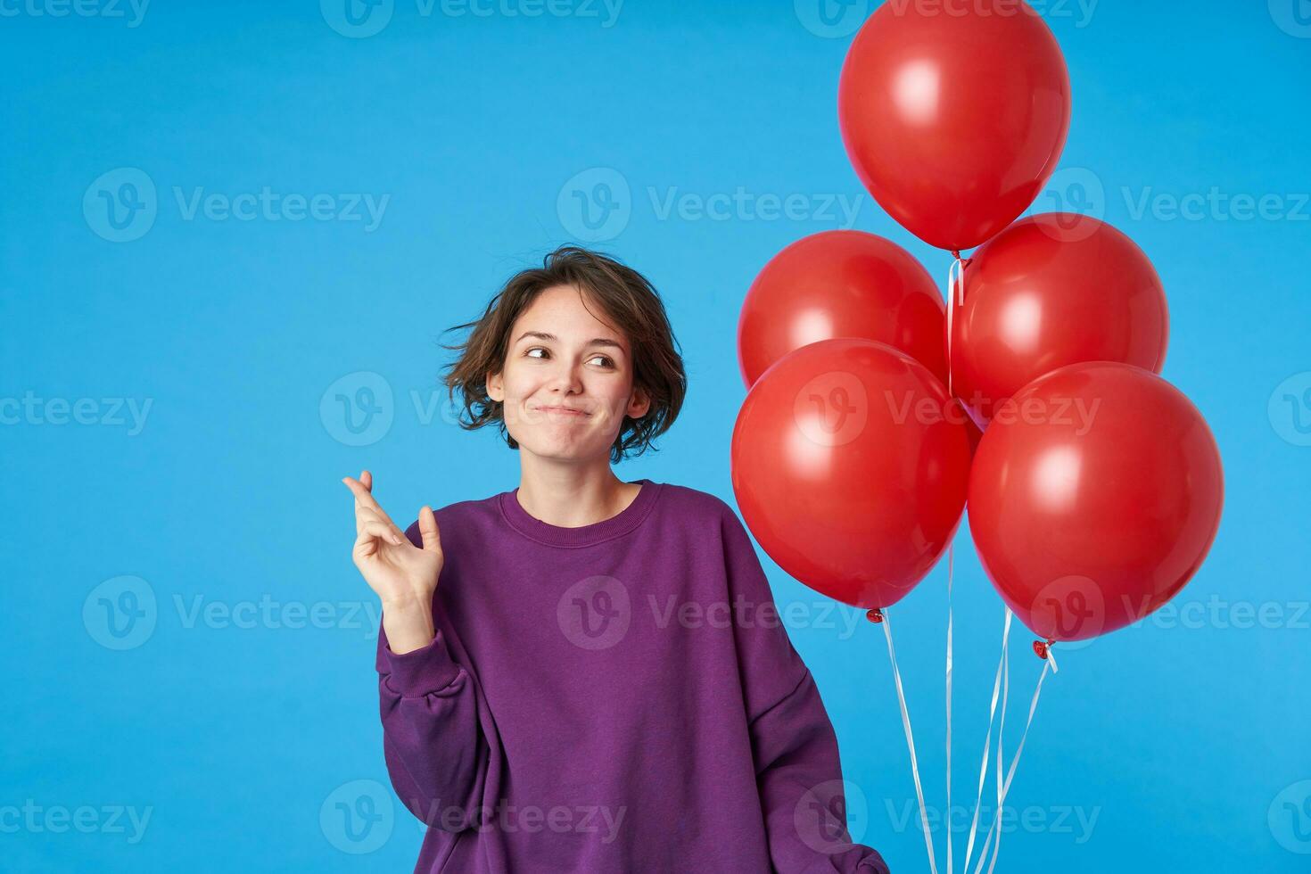 Indoor photo of young lovely curly brunette female with natural makeup making wish on her birthday and raising hands with crossed fingers, isolated over blue background