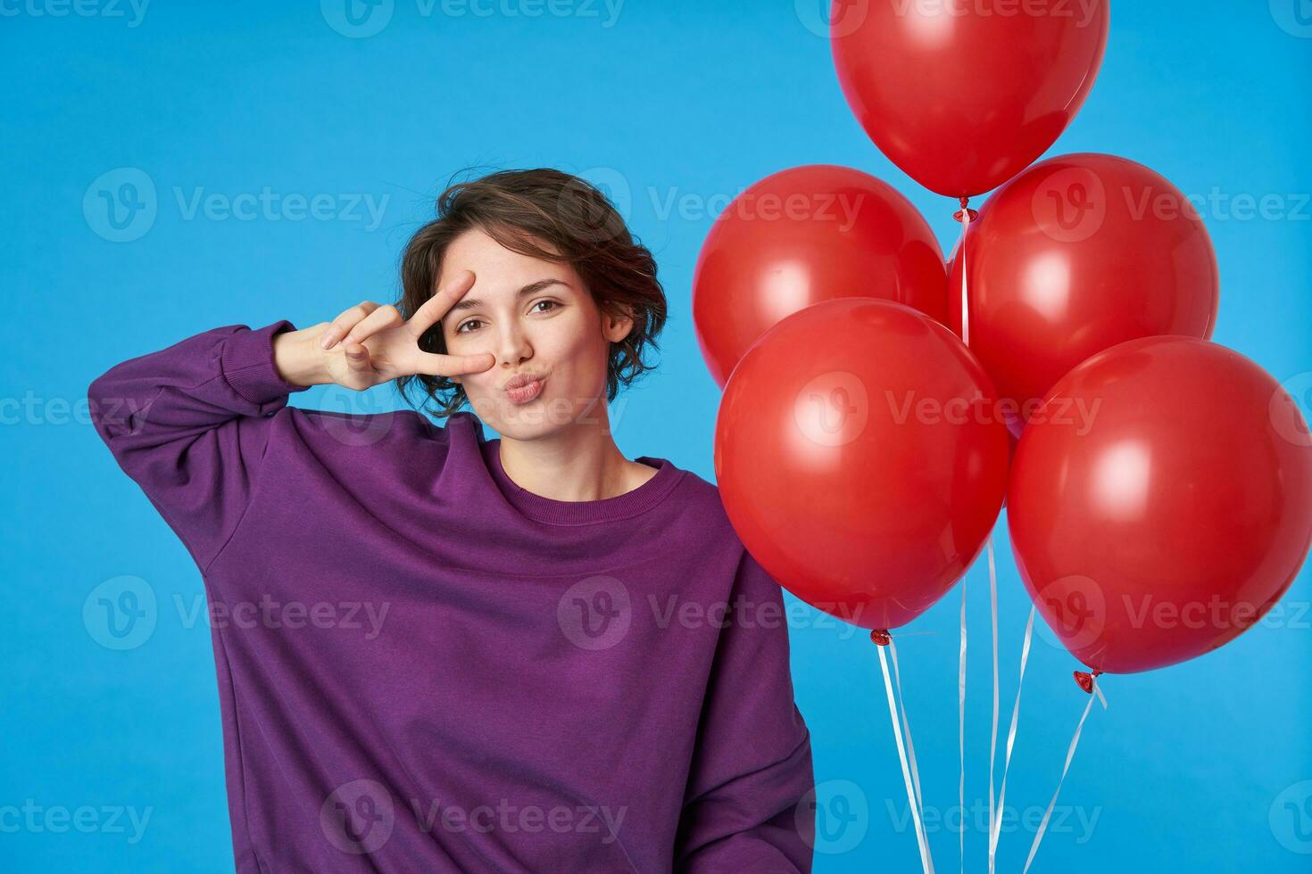 Glad young attractive curly lady with casual hairstyle folding her lips and raising to her face hand with peace gesture, dressed purple sweatshirt while posing over blue background photo