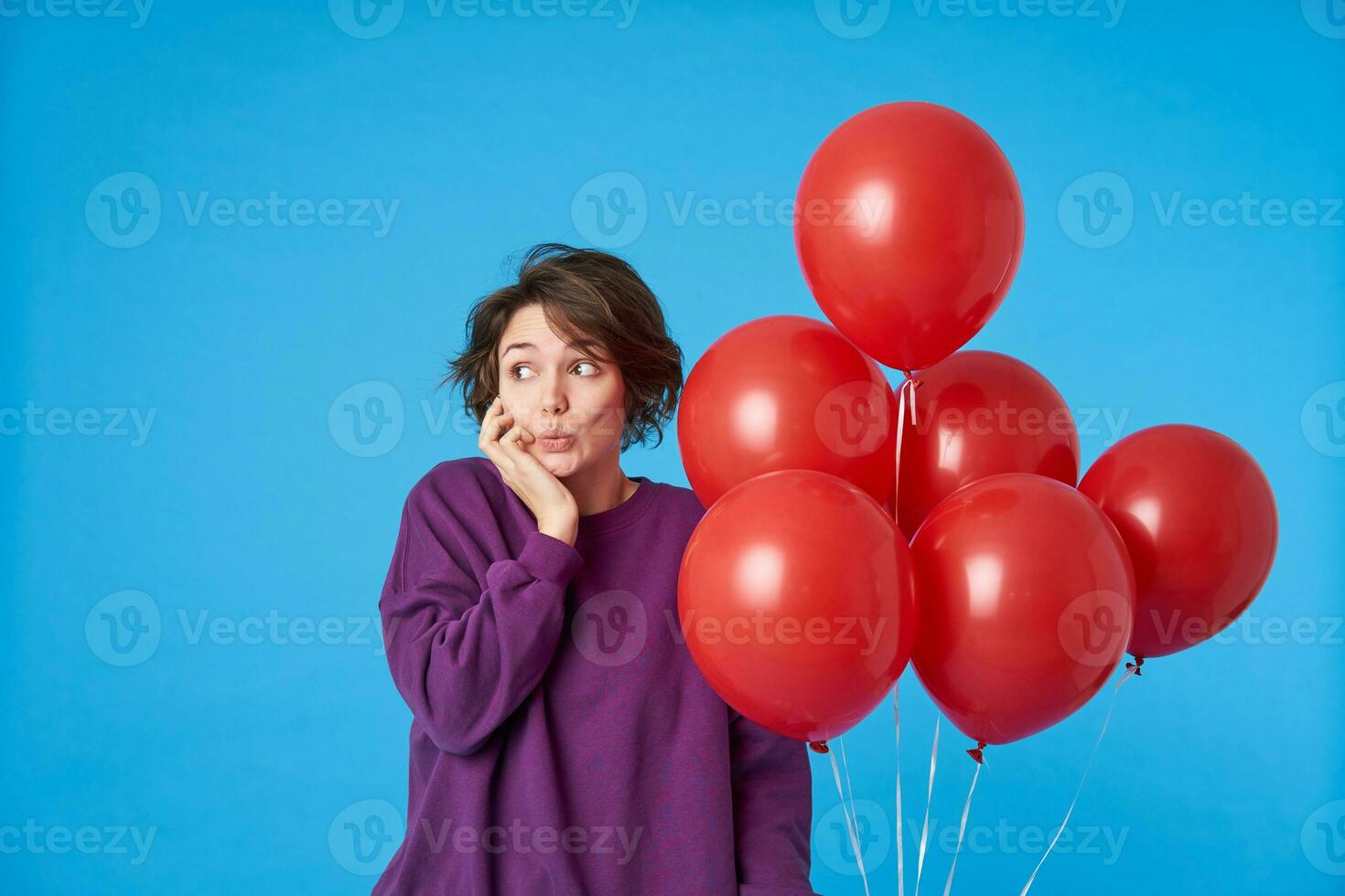 Positive young attractive dark haired female with trendy haircut folding her lips while looking surprisedly aside with raised eyebrows, standing over blue background photo