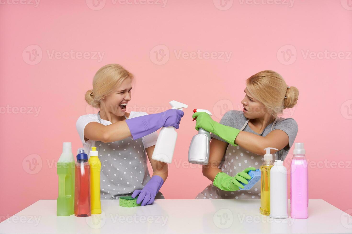 Studio photo of young white-headed cleaning ladies dressed in basic t-shirts and aprons raising hands with household chemicals and looking aexcitedly on each other, isolated over pink background