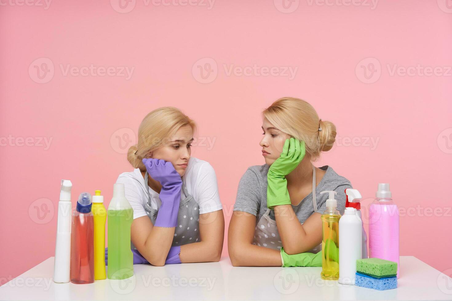 Tired young white-headed ladies with bun hairstyle leaning their exhausted faces on raised hands, having hard working day and relaxing over pink background after it photo