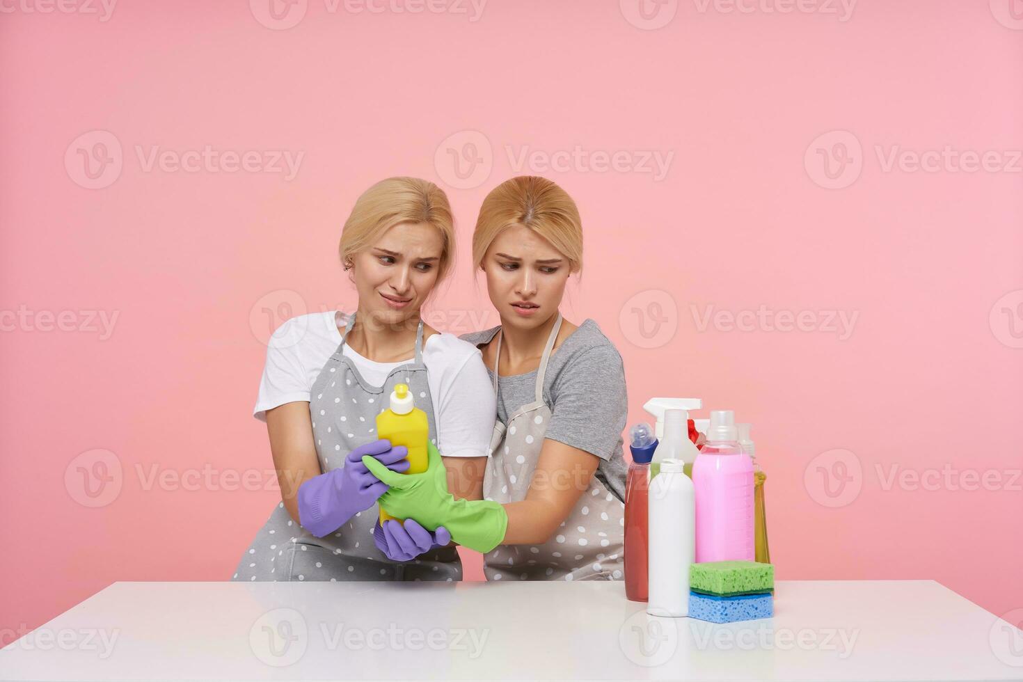 Confused young blonde women with natural makeup frowning their faces while holding bottle of detergent, sitting over pink background in basic t-shirts and aprons with dots photo