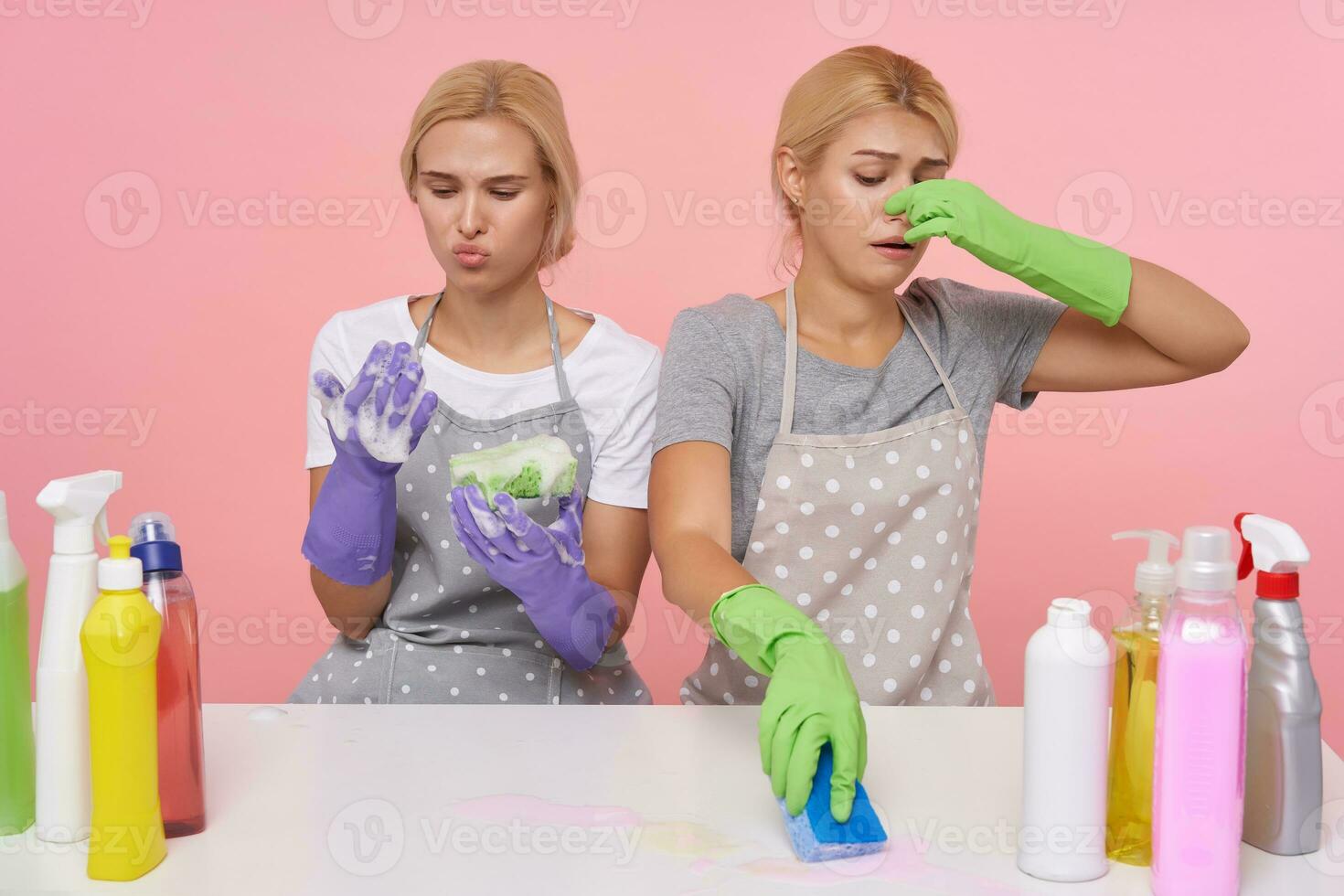 Displeased young blonde cleaning ladies with casual hairstyle wiping out stinky stain with detergents and sponges, sitting over pink background in working clothes and rubber gloves photo