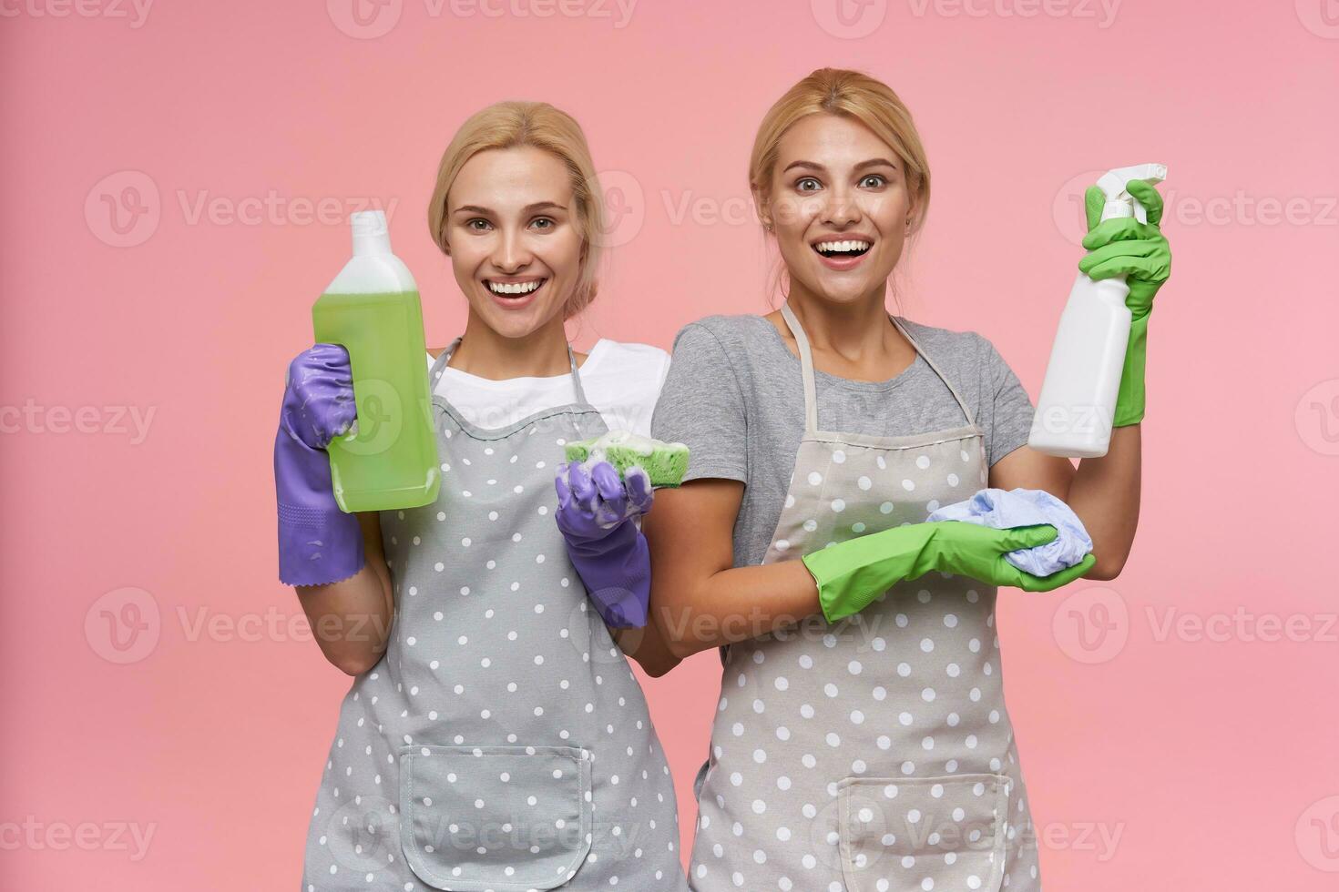 Joyful young white-headed cleaning ladies dressed in uniform keeping cleaning stuff in their hands while standing over pink background and smiling cheerfully photo