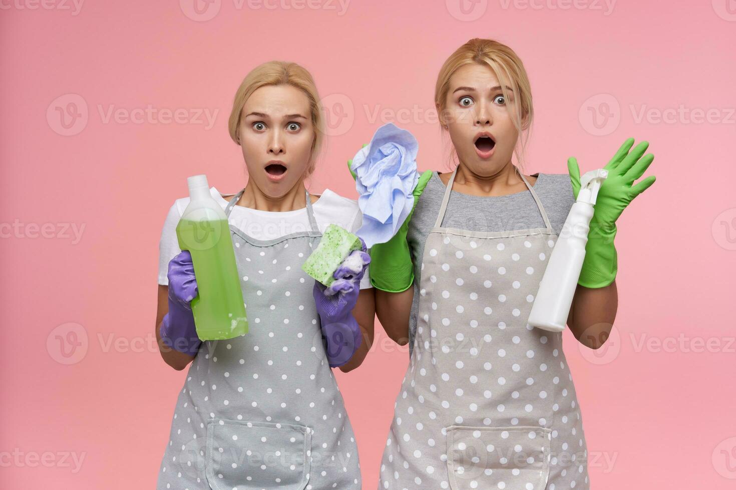Astonished young blonde women in rubber gloves looking at camera with wide eyes and mouth opened while standing over pink background with detergents in raised hands photo