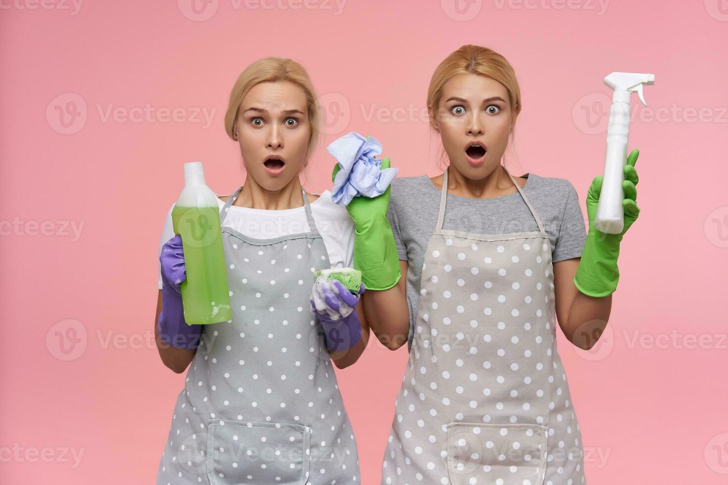 Dazed young pretty blonde cleaning ladies wearing casual t-shirts and polka dot apron looking amazedly at camera with opened mouths, isolated over pink background photo