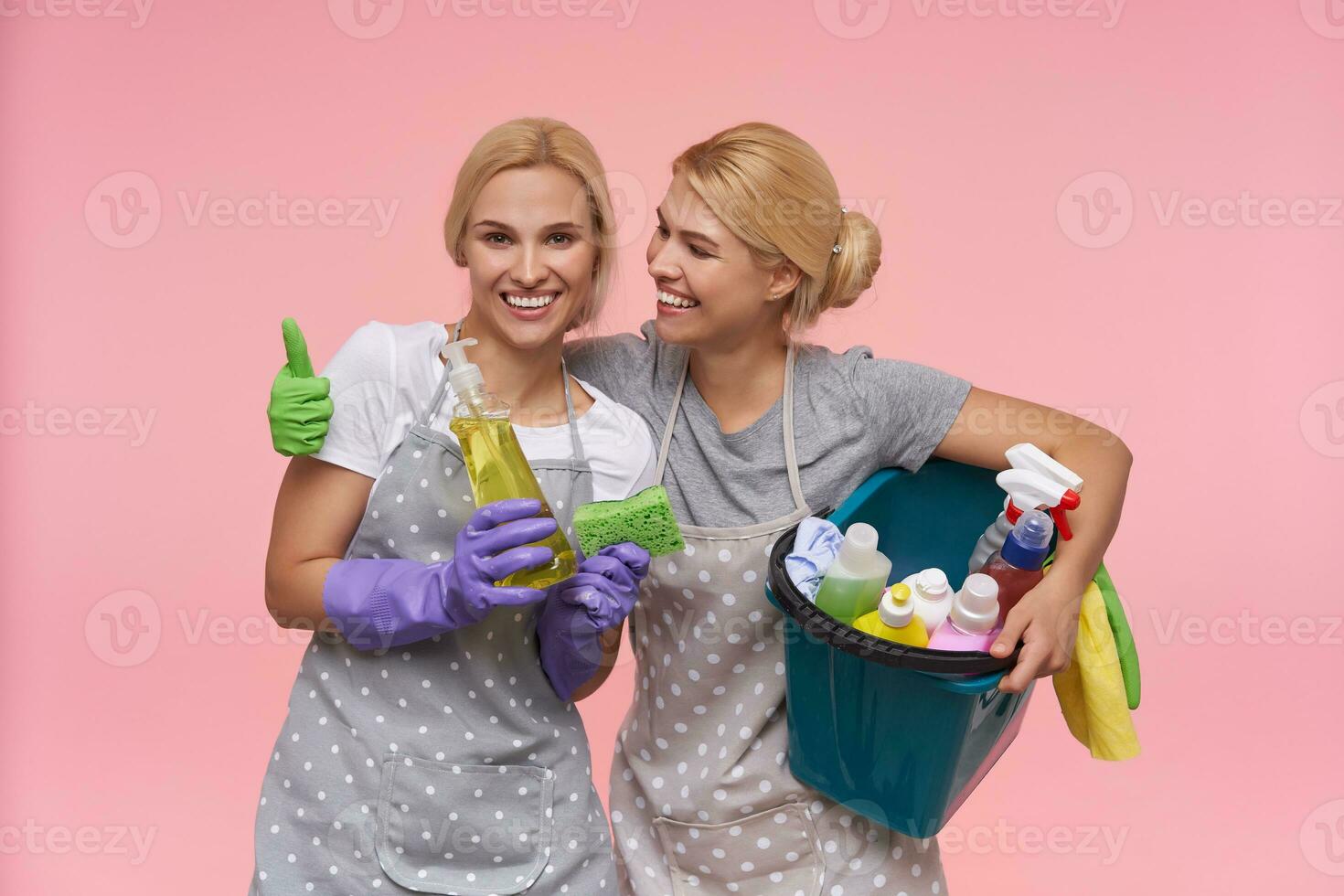 Joyful young white-headed housewives with bun hairstyle being in nice mood and smiling widely while standing over pink background in casual t-shirts and polka dot aprons photo