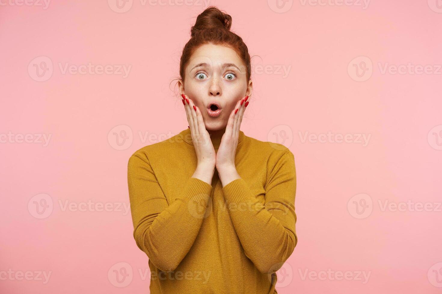 Amazed young pretty redhead woman with bun hairstyle holding her face with raised palms while looking at camera with wide eyes opened, isolated over pink background photo