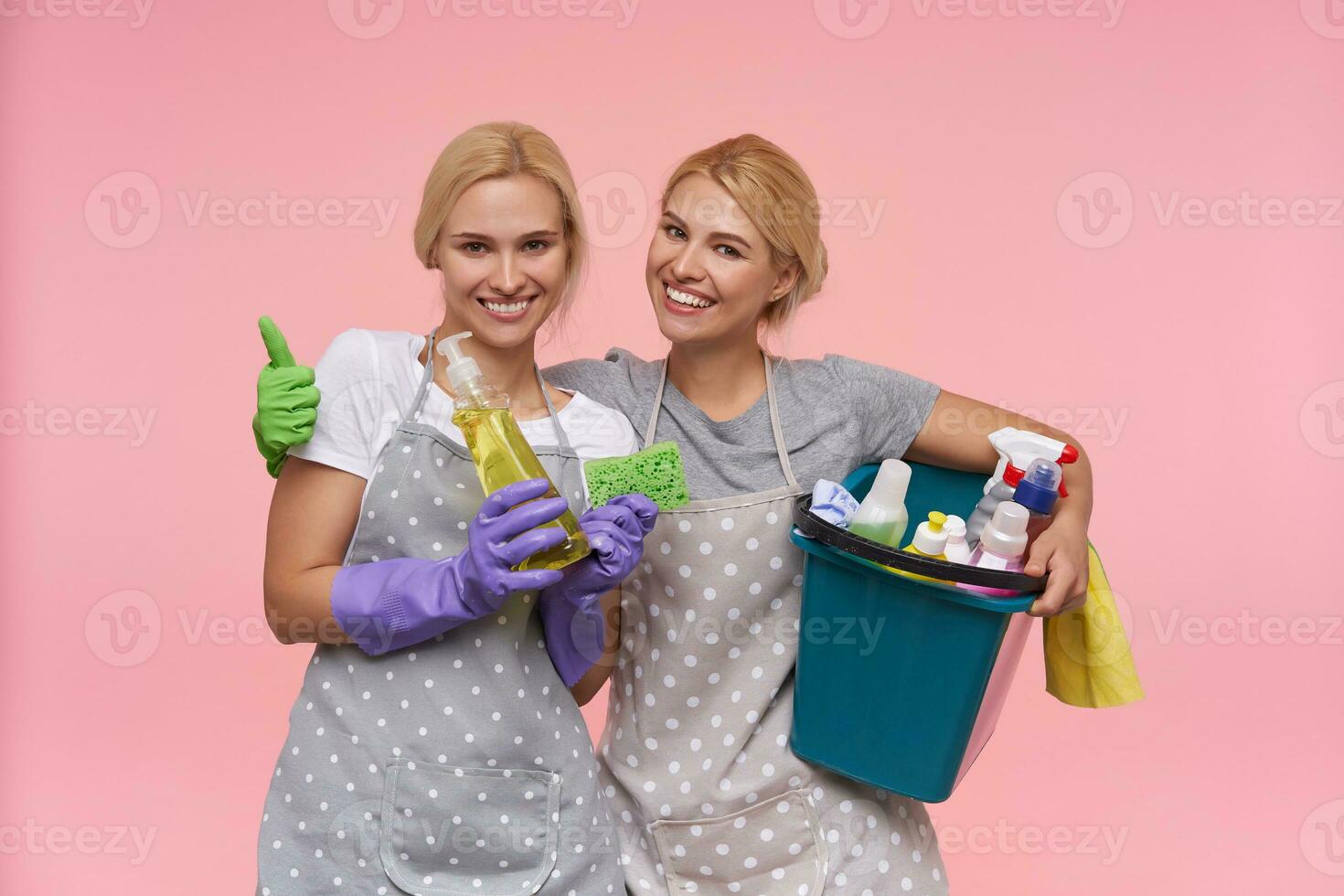 Glad young brown-eyed white-headed cleaning ladies dressed in working clothes and rubber gloves keeping cleaning stuff in their hands and smiling happily while finishing their working day photo