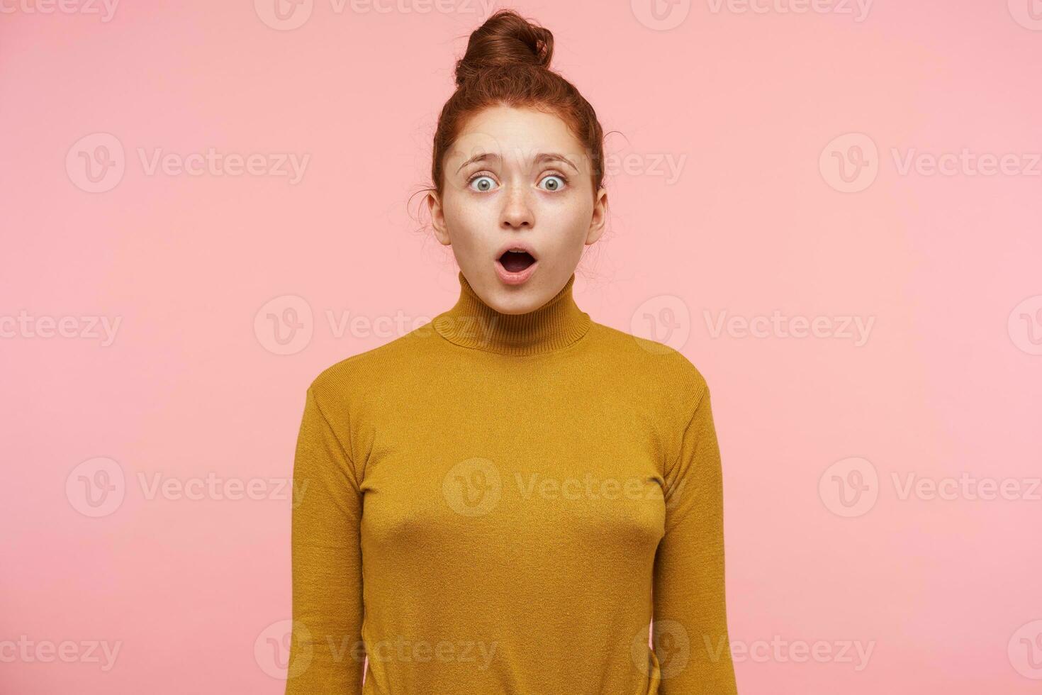 Indoor shot of shocked young attrative lady wearing her foxy hair in knot while standing over pink background, rounding her green eyes while lookng amazedly at camera photo