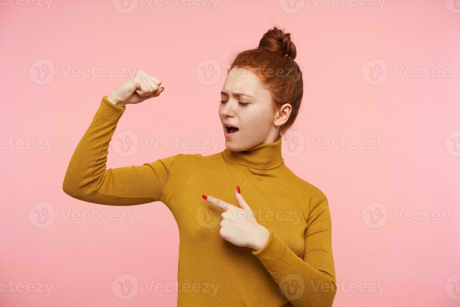 Young lady, self confident woman with ginger hair, freckles and bun. Wearing gold turtleneck sweater and pointing at her muscle on hand. Showing strength. Stand isolated over pastel pink background photo