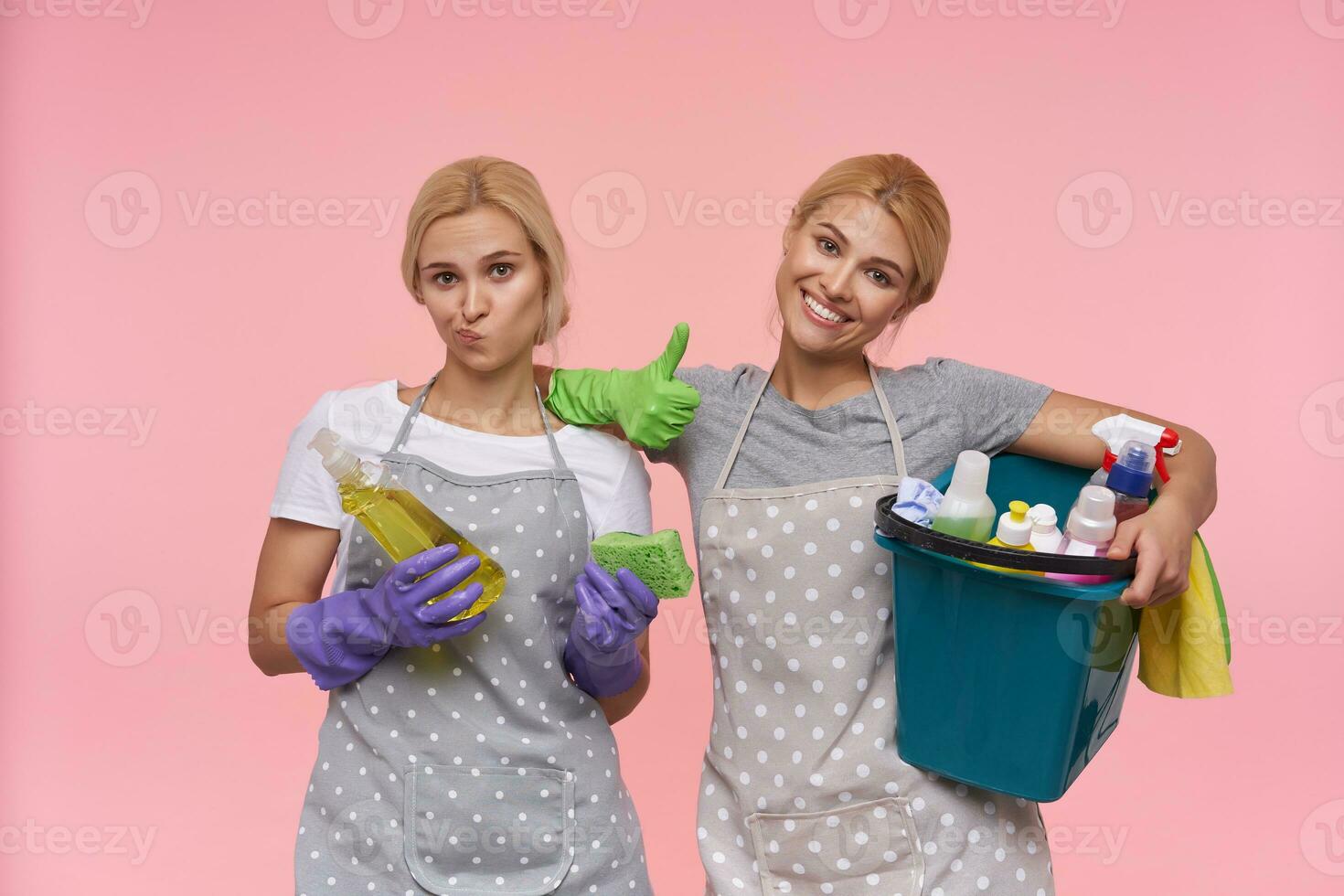 Happy young blonde cleaning lady showing raised thumb and smiling widely while her displeased colleague twisting her mouth with puzzled face, isolated over pink background photo
