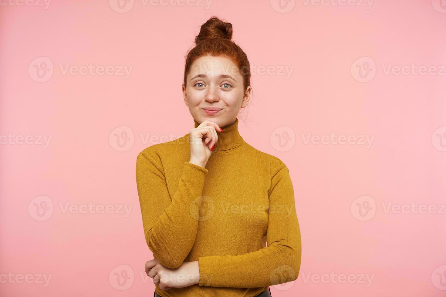 Portrait of attractive, adult girl with ginger hair, freckles and bun. Wearing gold turtleneck sweater and touching her chin, got an idea. Watching at the camera isolated over pastel pink background photo