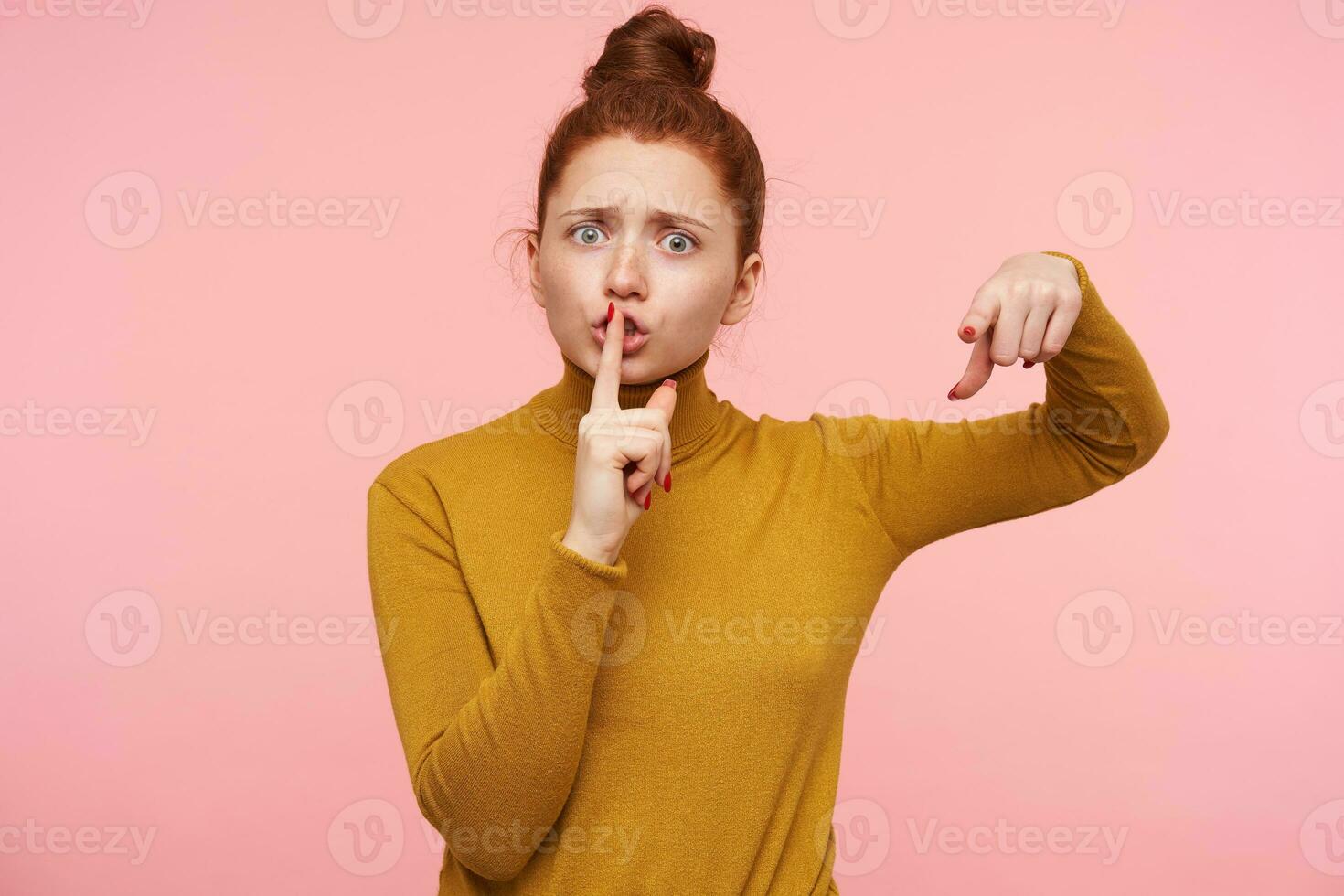 Severe young redhead pretty lady with casual hairstyle frowning her eyebrows while looking seriously at camera and raising hand with hush gesture, isolated over pink background photo