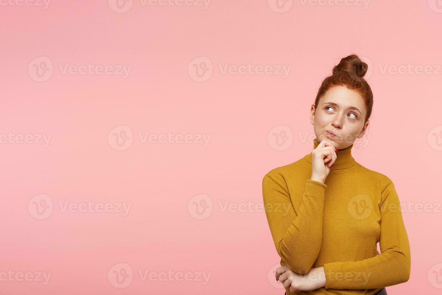Young lady, wonder woman with ginger hair, freckles and bun. Wearing gold turtleneck sweater and touching her chin in thoughts. Watching to the left at copy space over pastel pink background photo
