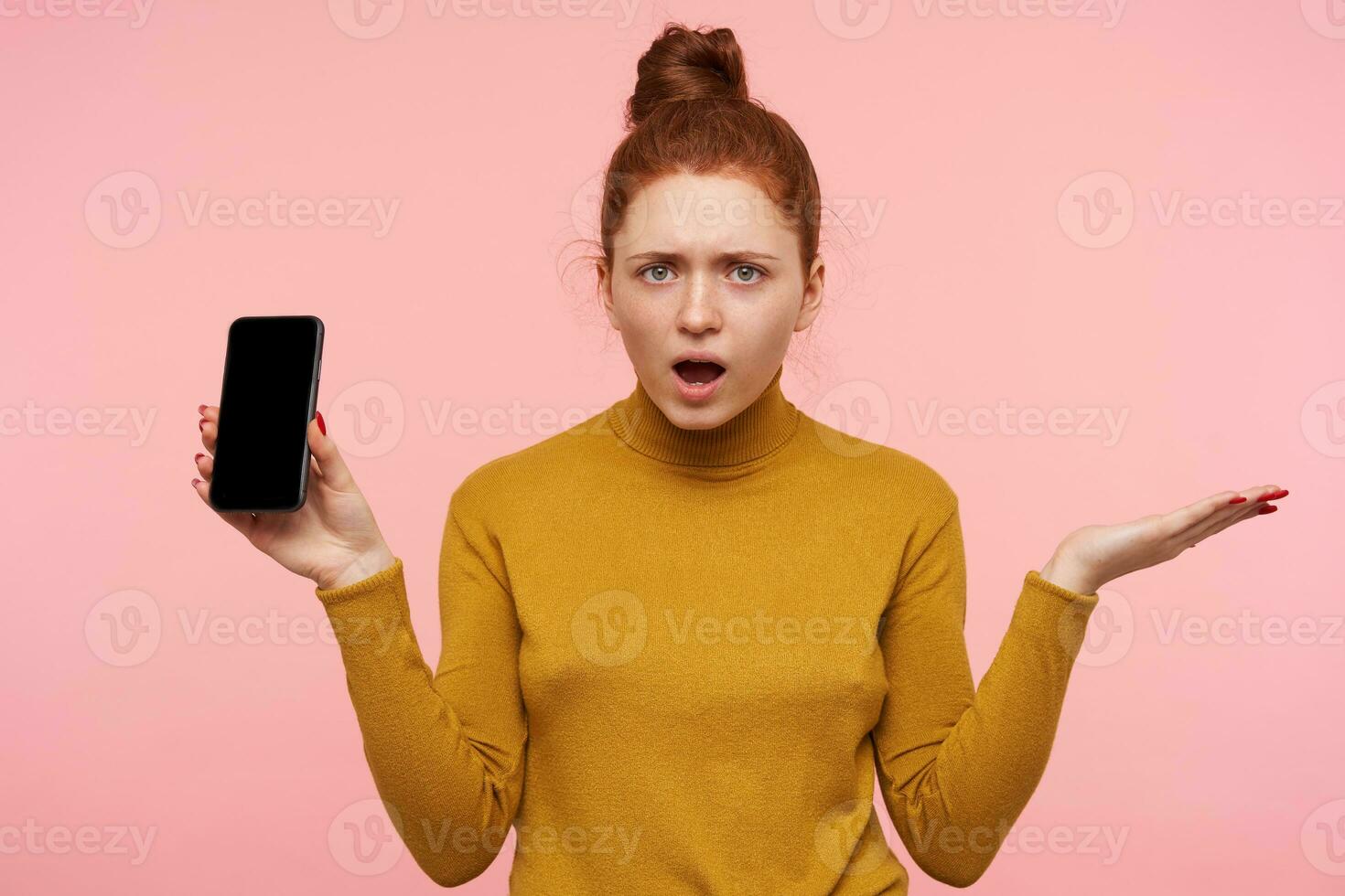 Studio photo of green-eyed young displeased redhead female raising perplexedly palm and frowning eyebrows while looking confusedly at camera, isolated over pink background
