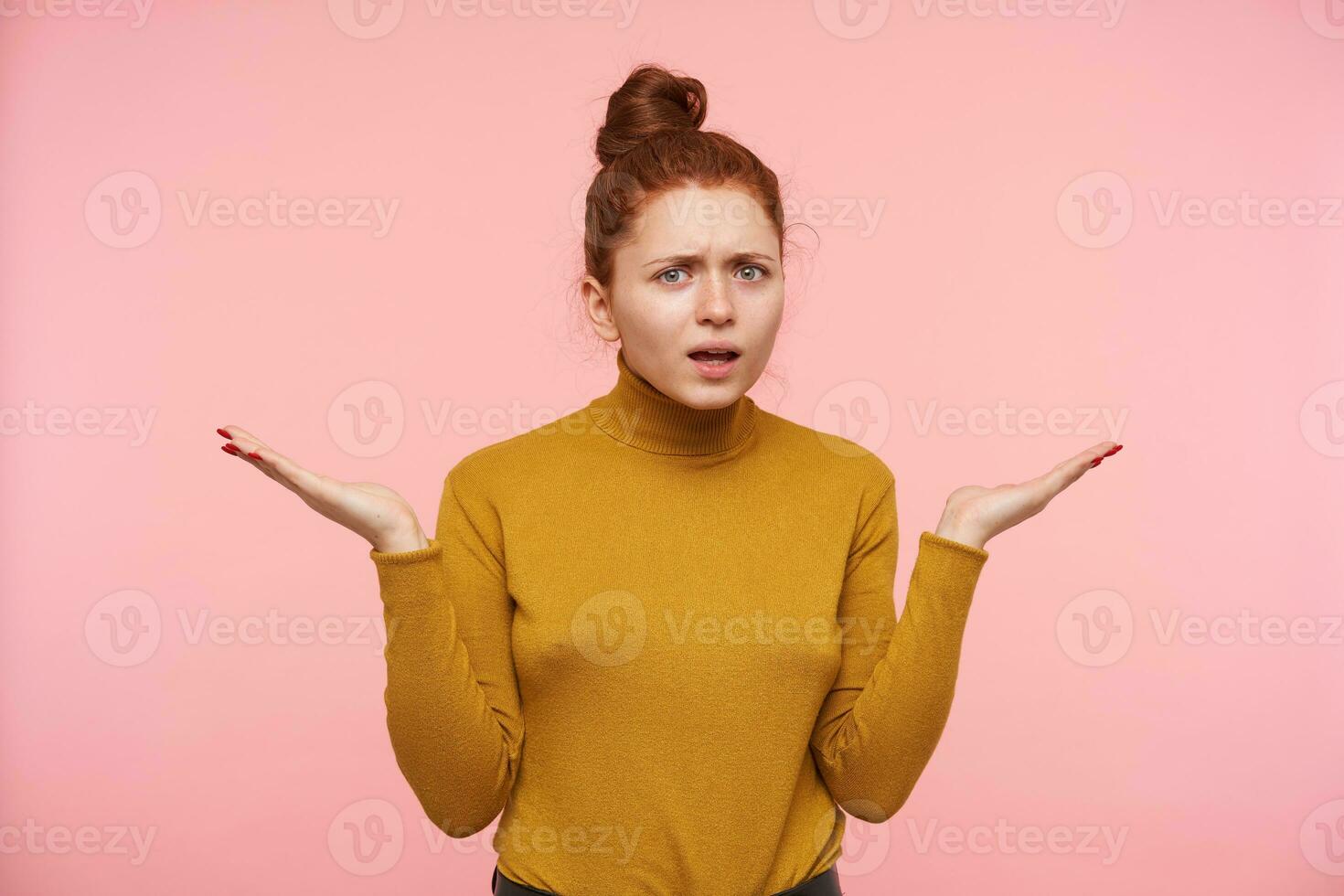 Puzzled young green-eyed redhead lady with natural makeup frowning her eyebrows and raising confusedly palms while looking at camera, standing over pink background photo