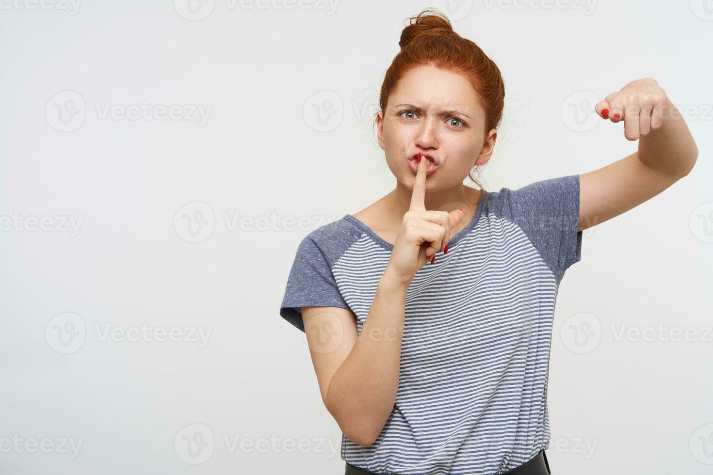 Severe young redhead lady with natural makeup frowning her eyebrows and asking to keep silence, holding index finger on her mouth while posing over pink background photo