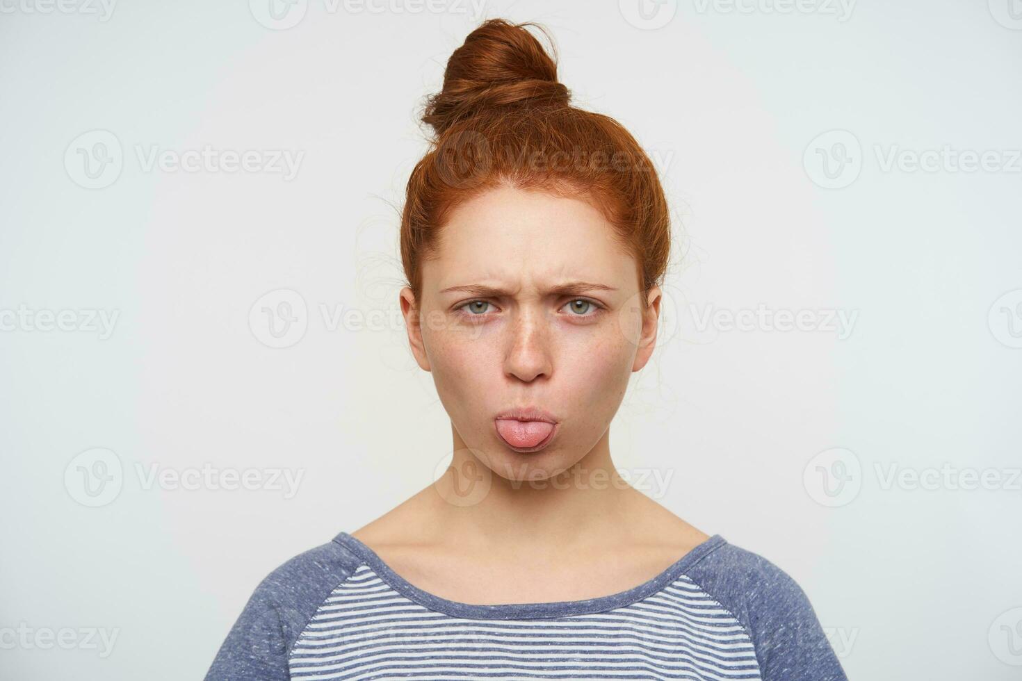 Offended young green-eyed redhead female frowning her eyebrows and showing tongue while looking sadly at camera, standing against pink background in blue t-shirt photo
