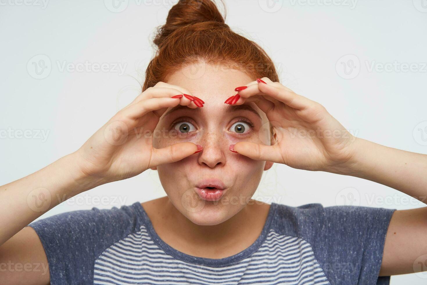 Close-up of excited young redhead female with red manicure looking at camera with wide eyes opened and fooling while standing over pink background in casual wear photo