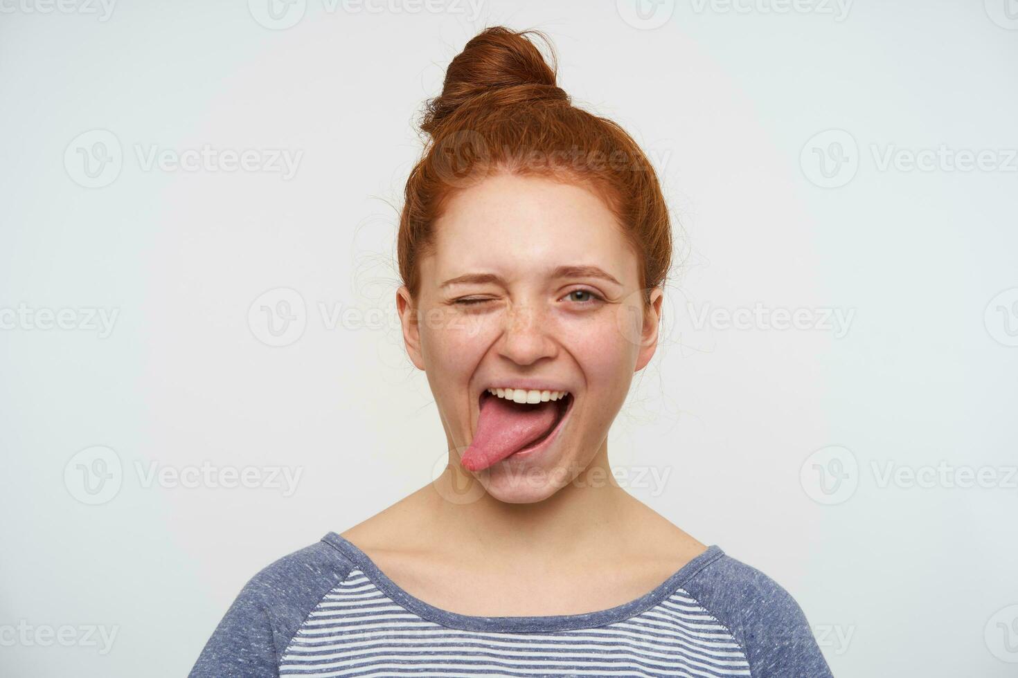 Joyful young loely redhead woman with natural makeup winking at camera and sticking happily out her tongue, having fun while posing over pink background photo