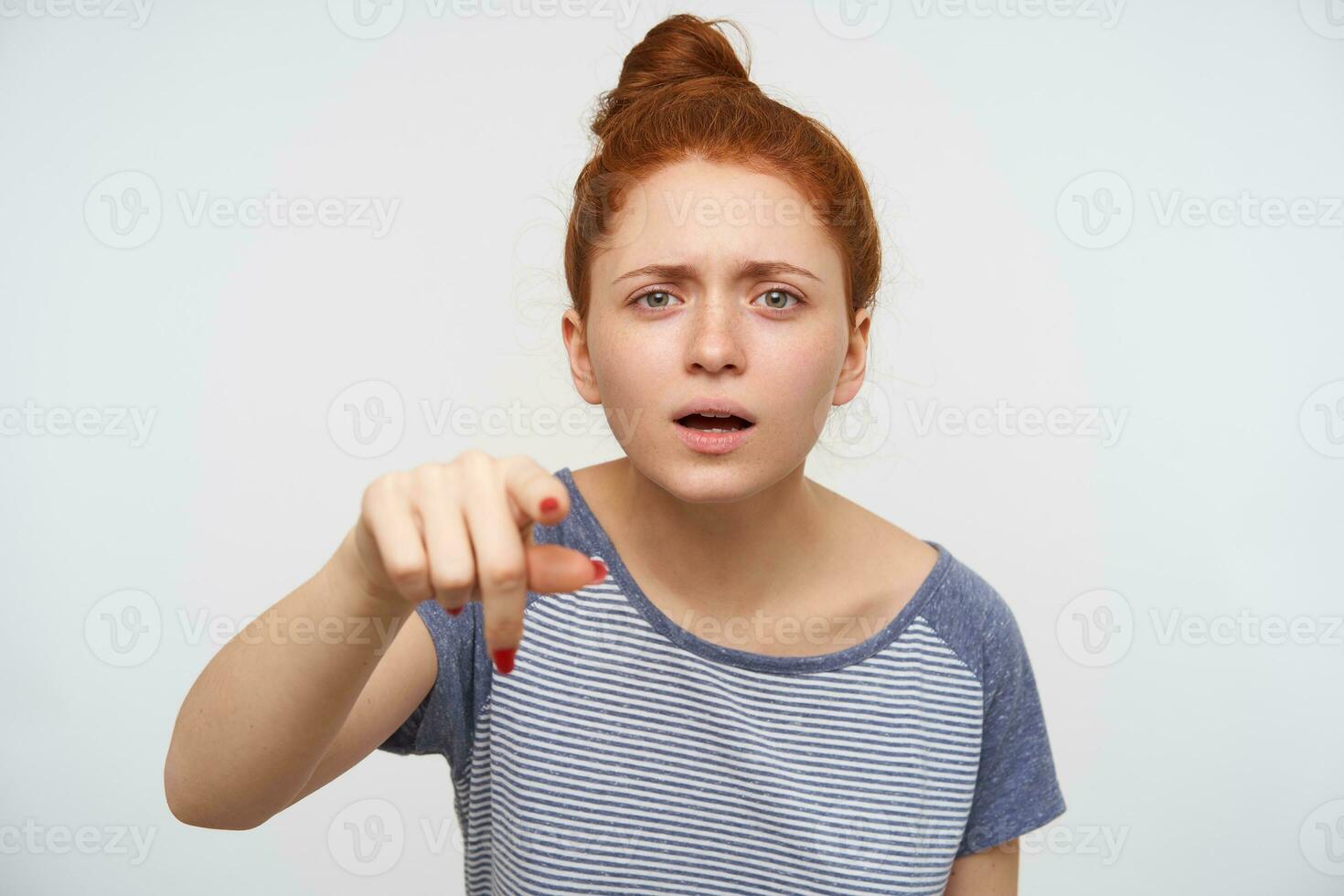 Puzzled young attractive redhead lady frowning her eyebrows while looking at camera and pointing ahead with raised forefinger, isolated over pink background photo