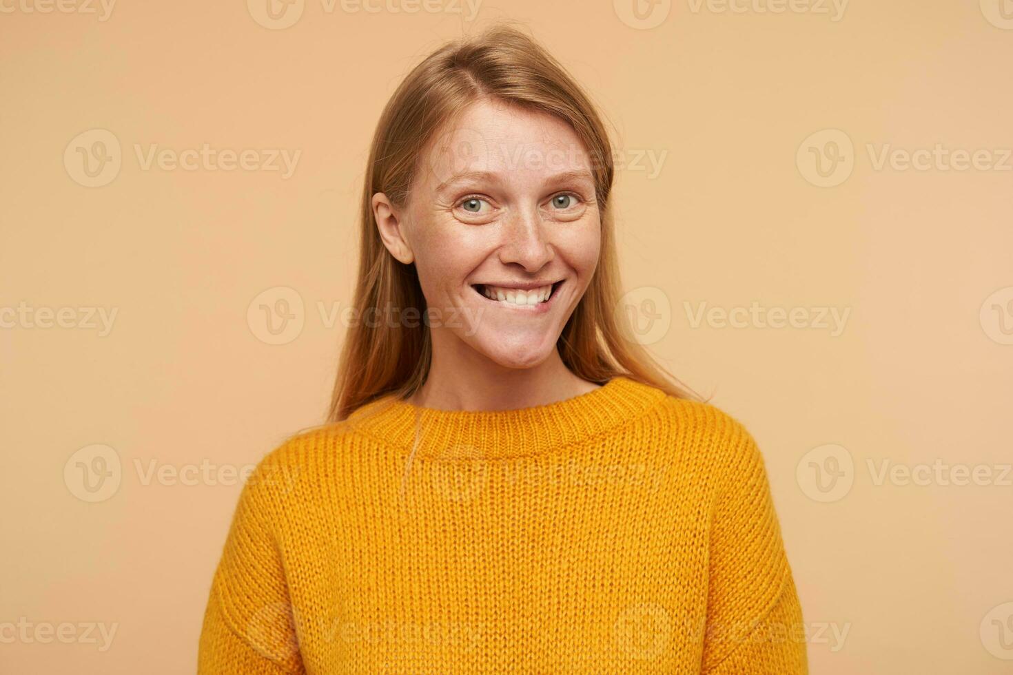 Portrait of young attractive glad lady with loose foxy hair looking at camera with charming smile and biting her underlip, standing against beige background photo