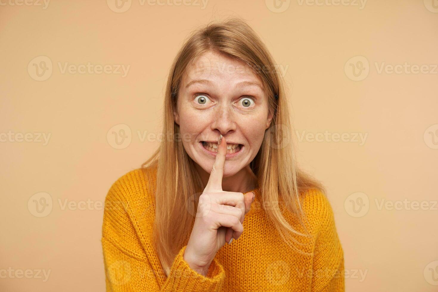 Indoor photo of young amazed redhead lady rounding her green eyes and holding raised forefinger on mouth while asking to keep secret, isolated over beige background