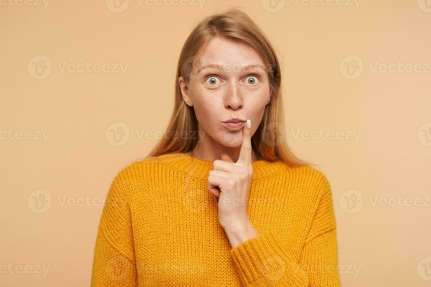 Bemused young lovely redhead woman rounding surprisedly her green eyes and holding raised forefinger on her cheek while being isolated over beige background photo