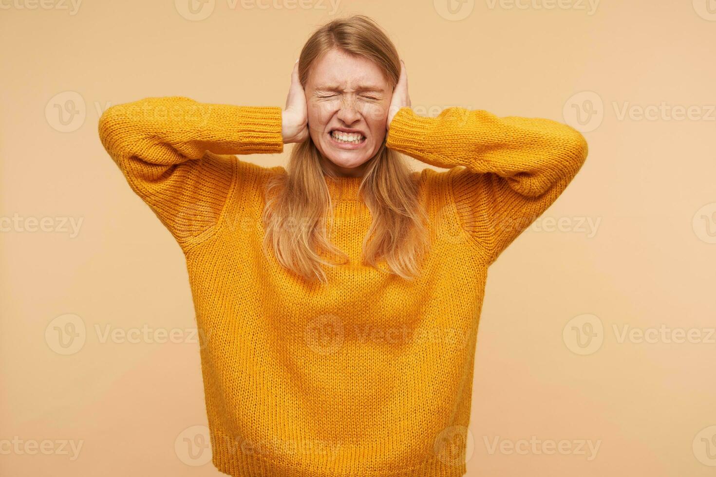 Stressed young redhead attractive woman frowning her face with closed eyes and closing ears with raised hands while avoiding loud sounds, standing over beige background photo