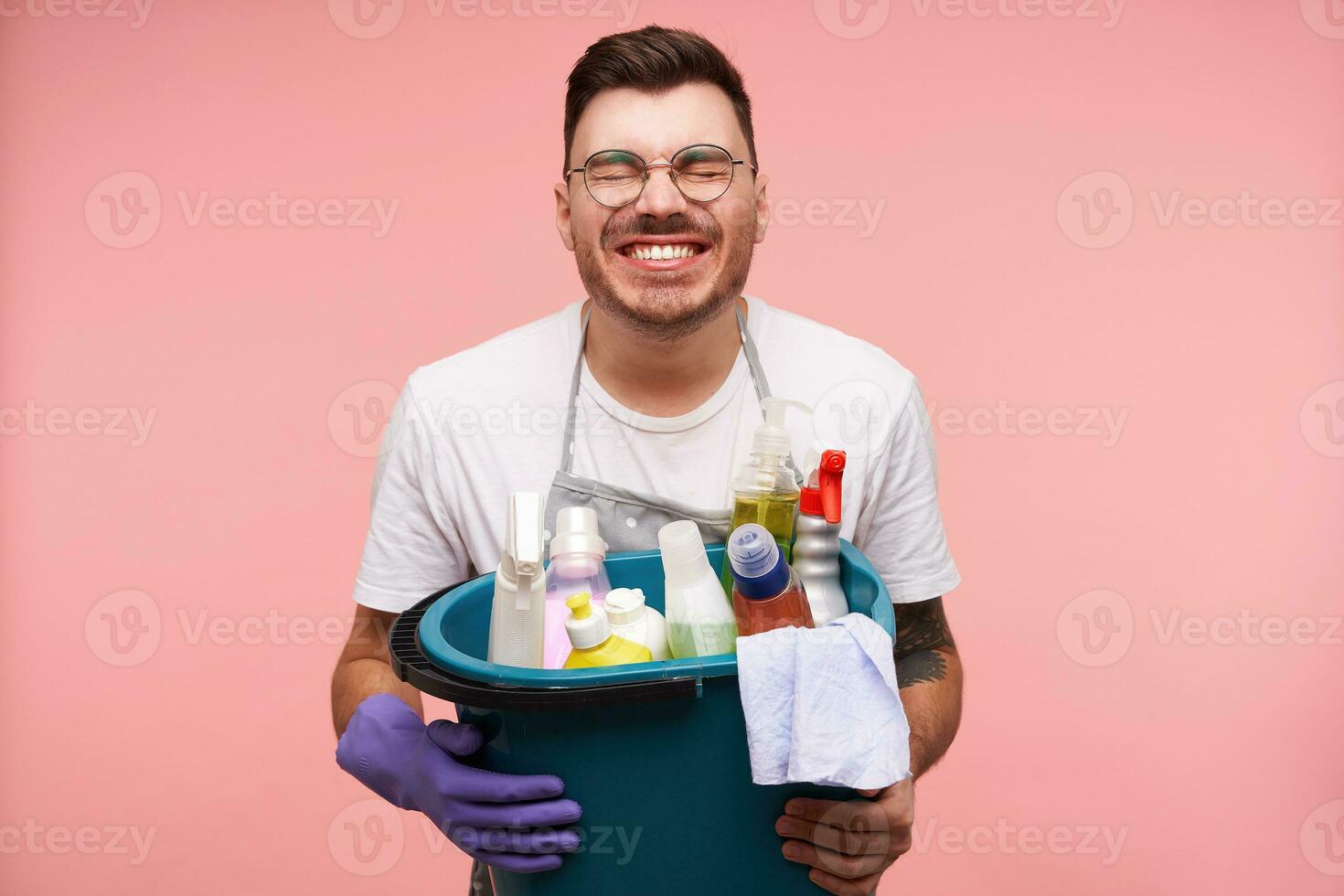 Cheerful young good looking brunette male in eyeglasses keeping his eyes closed and smiling happily while holding cleaning stuff, isolated over pink background photo