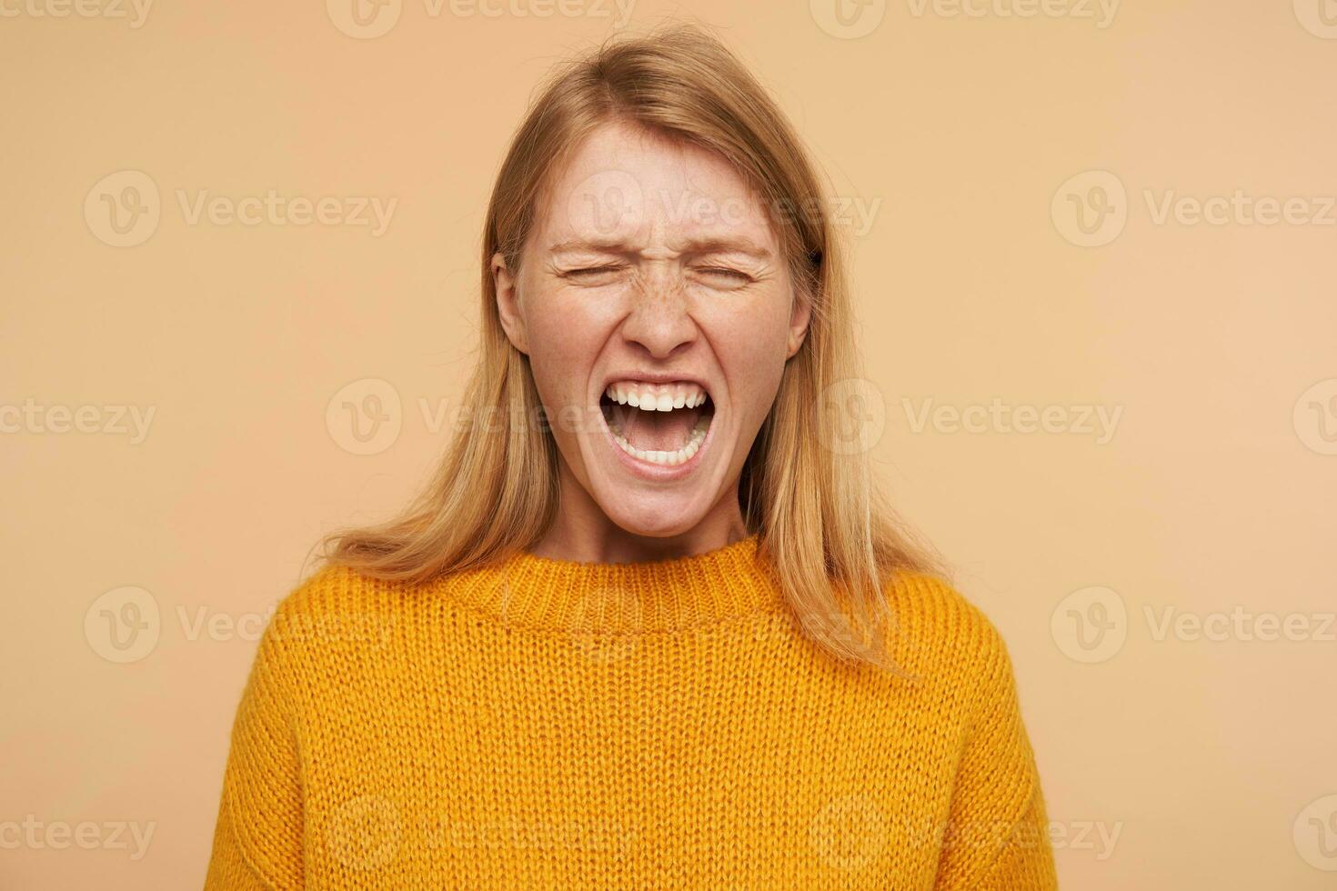 Stressed young long haired redhead female with casual hairstyle keeping her eyes closed while grimacing crossly her face, isolated over beige background photo
