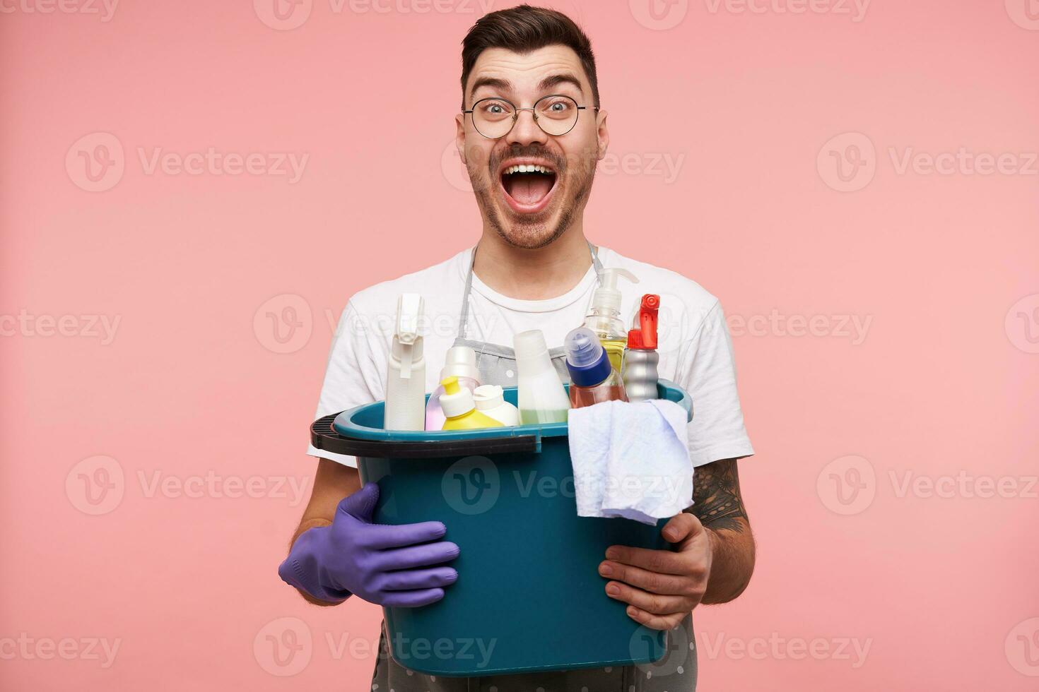 Excited young brunette bearded man with short haircut looking at camera with wide eyes and mouth opened while holding detergents, standing over pink background photo