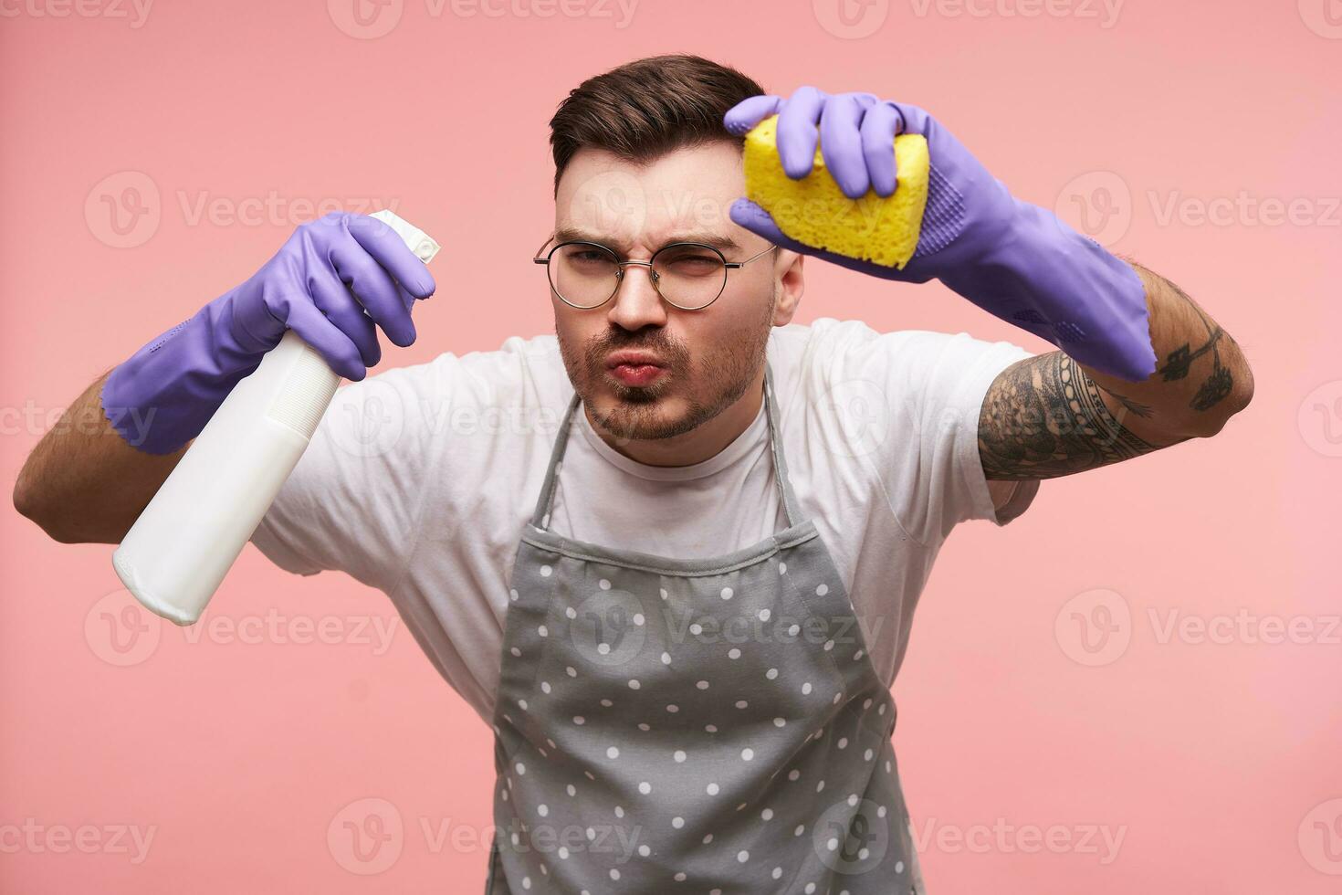 retrato de tatuado joven perplejo morena hombre con corto Corte de pelo persiguiendo su labios y bizco ojos mientras posando terminado rosado antecedentes en delantal, blanco camiseta y caucho guantes foto