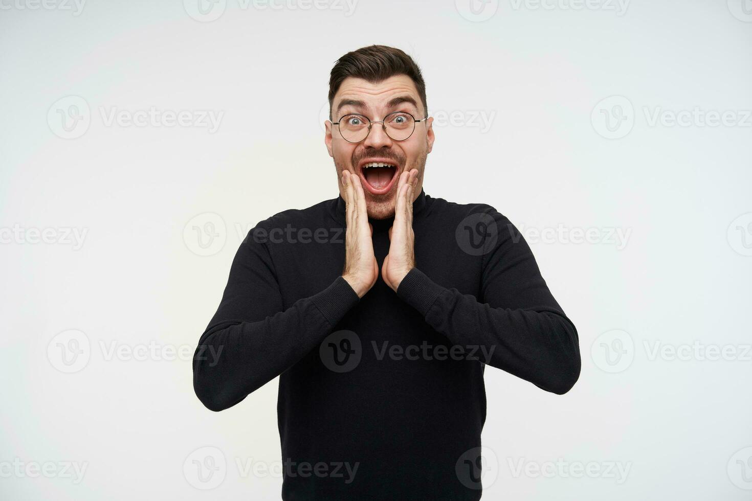 Bemused young pretty dark haired bearded guy holding palms on his face while looking amazedly at camera with wide eyes and mouth opened, isolated over white background photo
