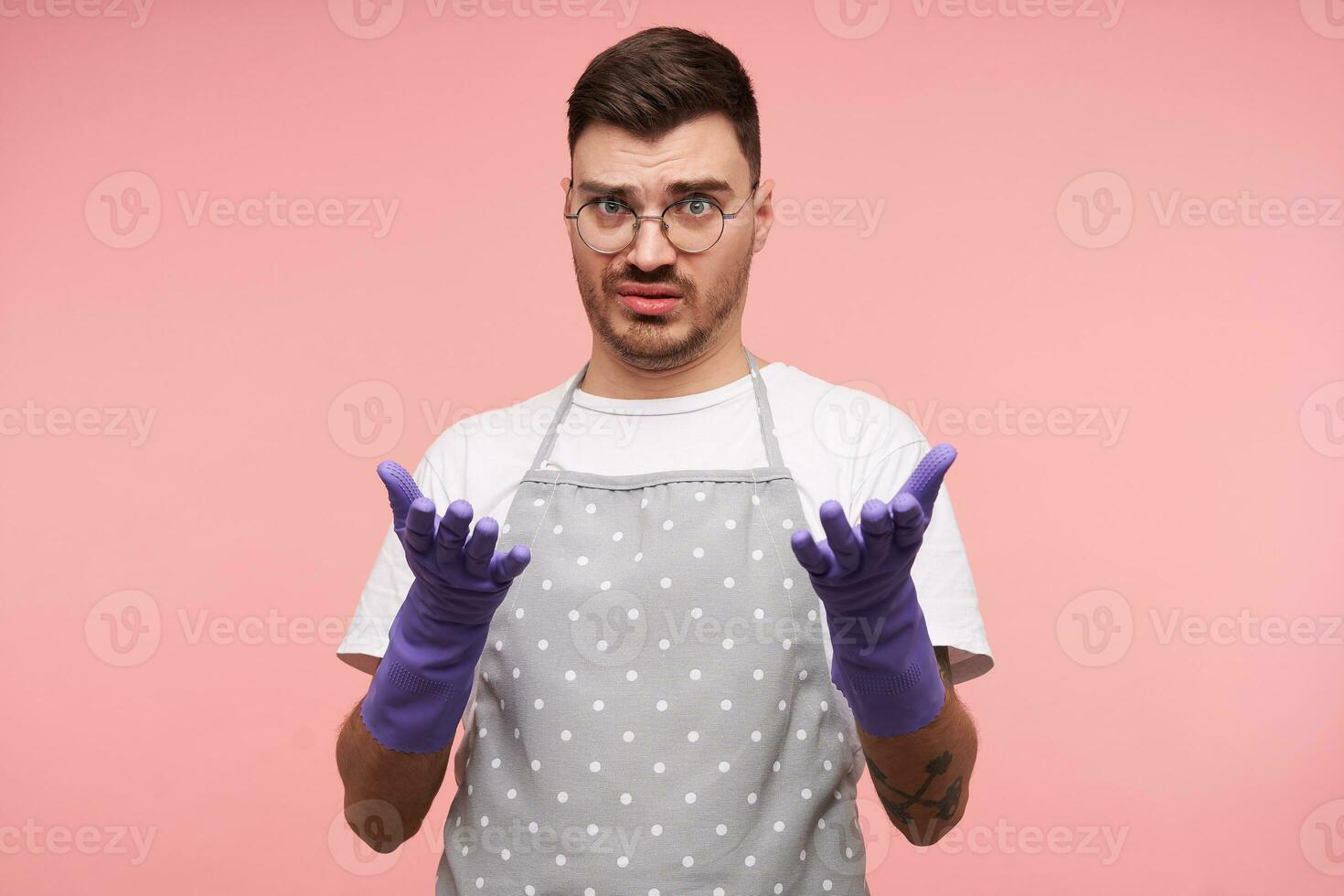 Bewildered young brown haired male with short haircut looking confusedly at camera with raised hands while standing over pink background in working clothes and rubber gloves photo