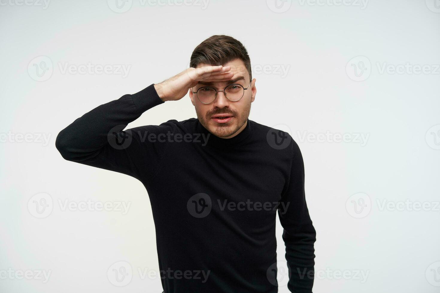 Portrait of young handsome dark haired man in eyewear squinting his eyes and keeping raised hand on forehead while trying to see something far away, posing over white background photo