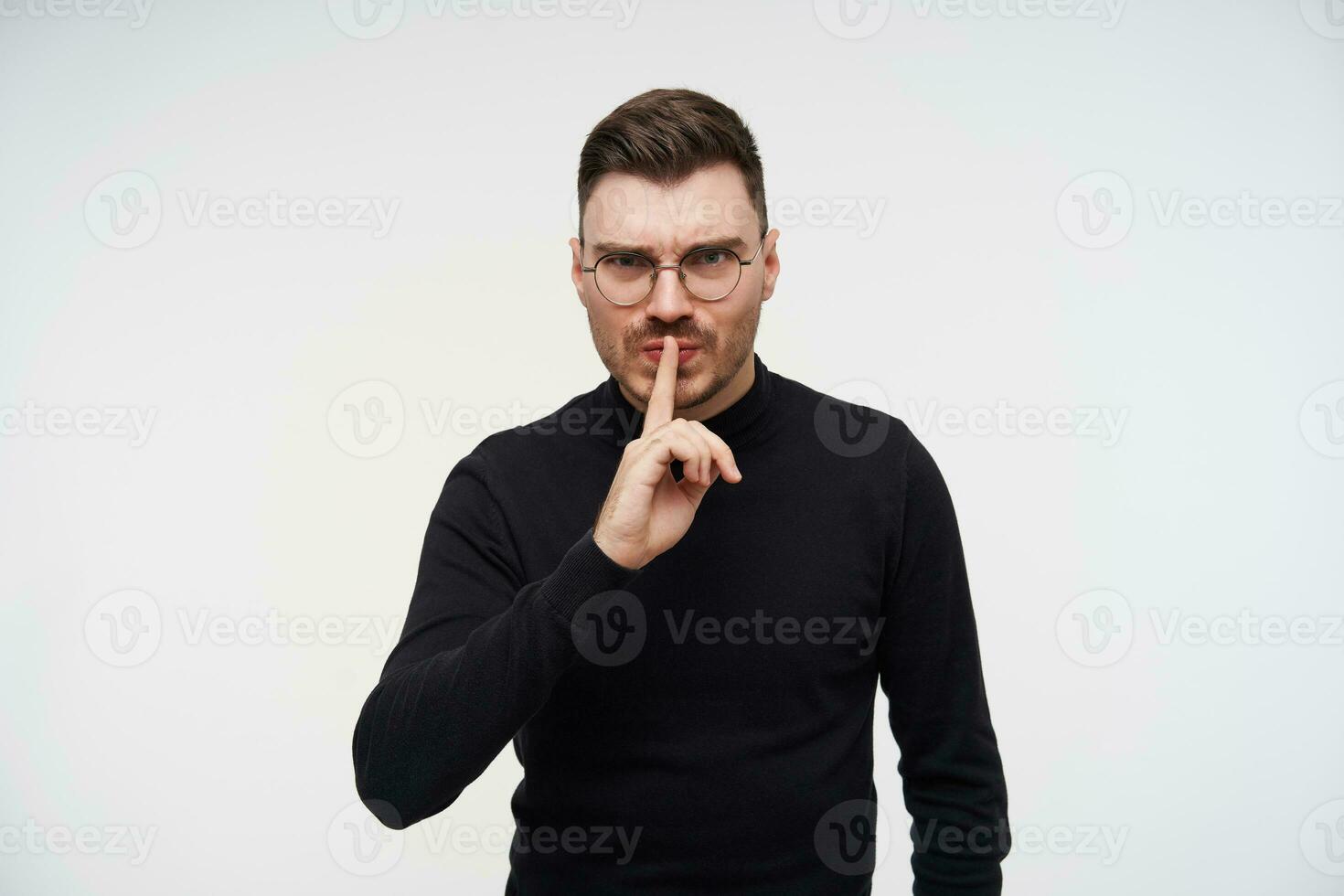 Severe young attractive dark haired male frowning his eyebrows while asking to keep silence and raising hand in hush gesture, posing over white background photo