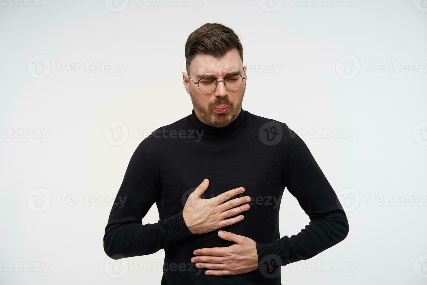 Indoor photo of young bearded brunette man with short haircut holding palms on his stomach and grimacing his face, feeling sick while posing over white background