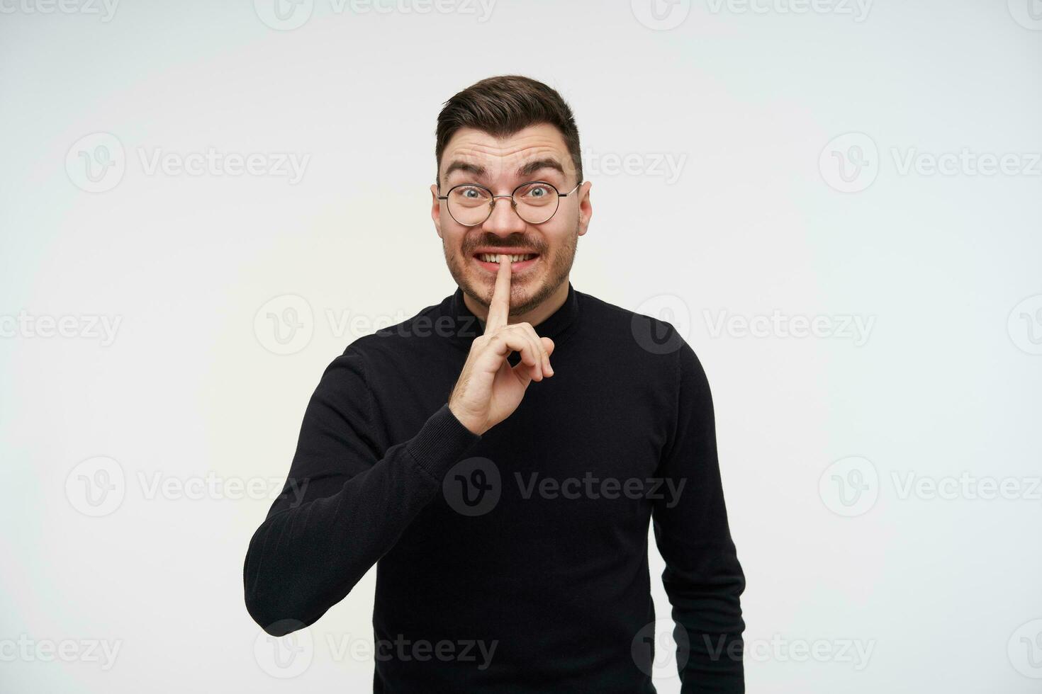 Portrait of excited young brown haired bearded man keeping raised forefinger on his mouth and raising surprisedly eyebrows while standing over white background photo