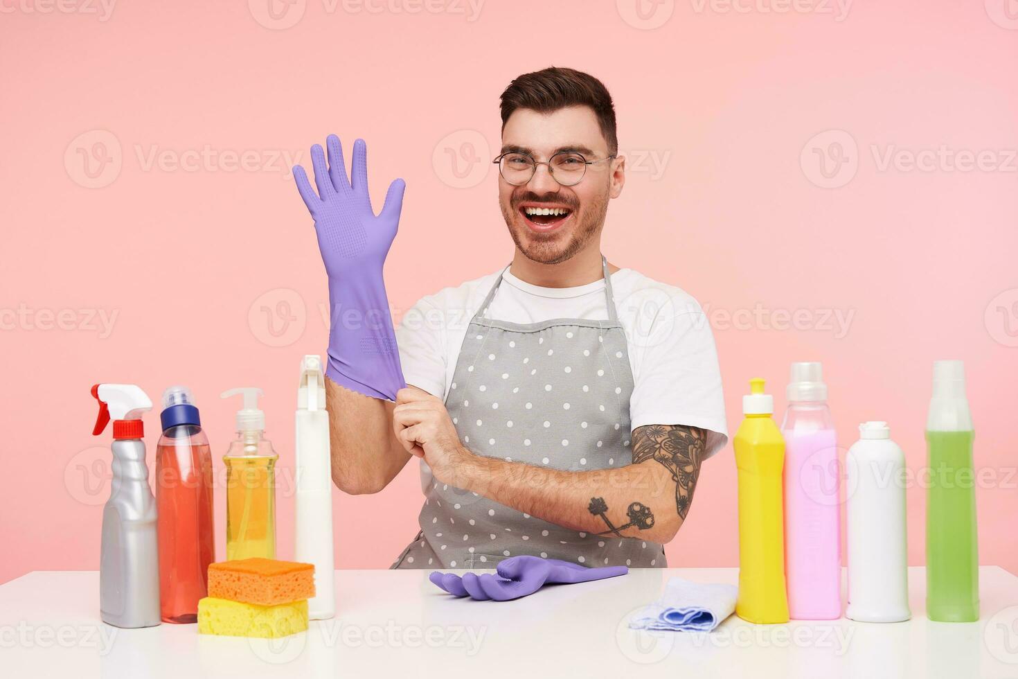 Cheerful young pretty bearded dark haired man preparing for spring cleaning and looking happily at camera, sitting over pink background with household chemicals photo